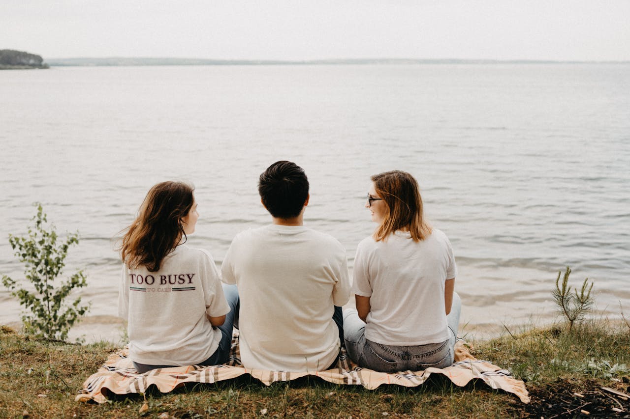 Three friends sitting on a blanket by the ocean, enjoying a peaceful beach picnic together.
