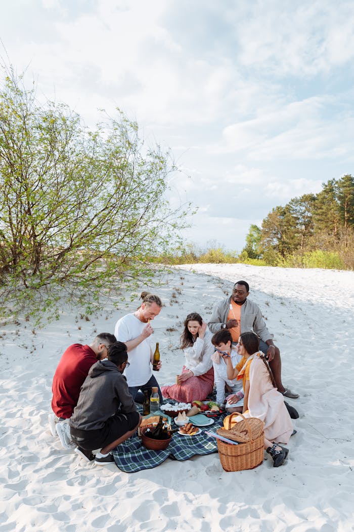 A diverse group of friends enjoying a picnic on a sandy beach with food and drinks.